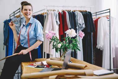 Düşük açılı, elinde gözlüklerle masada oturan ve rahat bir elbise atölyesinde çalışırken gülümseyen pozitif bir kadın.