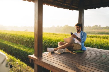 Tam vücut pozitifliği olan yazlık kıyafetli, bacak bacak üstüne atmış, akıllı telefonlu, uzaktan kumandalı, ahşap teras üzerinde çalışan yeşil çayır çevrili bir adam.