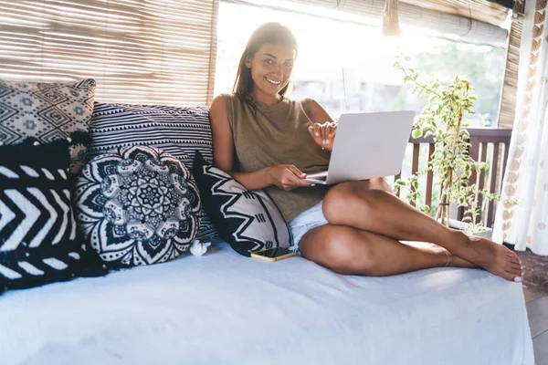 Tüm vücudu çıplak ayaklı, yatakta oturup verandada sörf yaparken kameraya bakan etnik bir kadın.