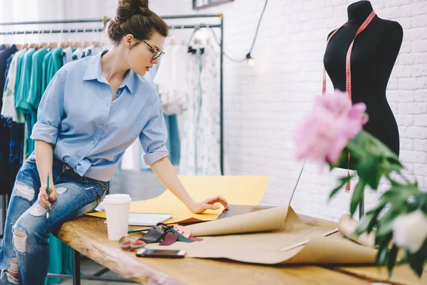 Modern atölyede çalışırken dizüstü bilgisayarını kullanarak kahve ve dikiş desenleriyle masada oturan düşünceli terzinin yan görüntüsü.