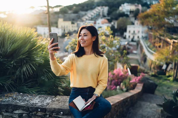 Kitapla gülümseyen ve parapet üzerinde otururken akıllı telefondan video görüşmesi yapan sıradan giyinmiş mutlu genç Asyalı kadın.