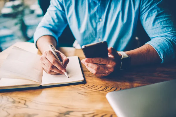 Modern akıllı telefonu tutan ve bilgi aramak için yüksek hızlı internet bağlantısı kullanarak not defterine metin mesajı yazan erkek ellerinin kırpılmış görüntüsü 