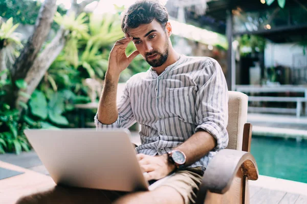 Kafası karışmış, sakallı genç bir adam. Düşünceli bir şekilde kaşlarını okşarken aynı zamanda dizüstü bilgisayarla proje üzerinde çalışıyor ve zor görevleri düşünüyor.
