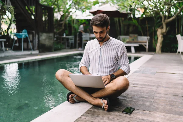 Young bearded male entrepreneur in summer shirt and flip flops working on laptop while sitting on floor near swimming pool in quiet backyard