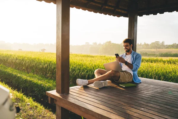 Tam vücut pozitifliği olan yazlık kıyafetli, bacak bacak üstüne atmış, akıllı telefonlu, uzaktan kumandalı, ahşap teras üzerinde çalışan yeşil çayır çevrili bir adam.