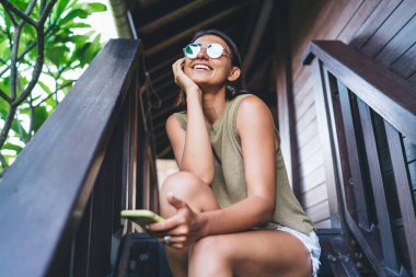 Düşük açılı, günlük kıyafetler ve güneş gözlüklü bir kadın tahta merdivenlerde oturuyor ve Bali 'de dinlenirken akıllı telefon sörfü yapıyor.