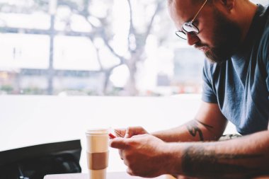 Tişörtlü, gözlüklü, elinde kahve bardağıyla oturmuş akıllı telefon kullanan ciddi bir erkeğin yan görüntüsü.