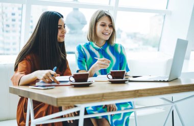 Genç çok ırklı bayan arkadaşlar yabancı dil öğreniyorlar. Dizüstü bilgisayar kullanıyorlar ve modern kafede sıcak içecekler içiyorlar.