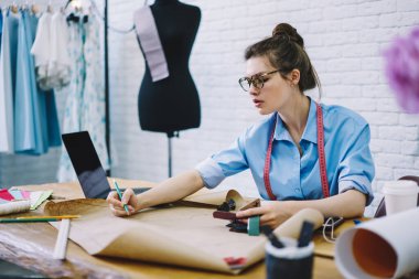 Gözlüklü konsantre kadın lağımı ve boynu boynundaki şerit çizimler, dizüstü bilgisayarlar ve kumaş örnekleriyle masada oturarak dikiş deseni yapmakla meşgul.