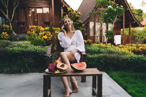 Full body of smiling female tourist sitting on table with fruits and looking away while talking via smartphone and chilling in resort