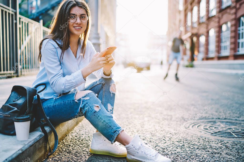 Portrait of brunette millennial student in stylish wear and eyewear posing in city during sunny day holding mobile phone