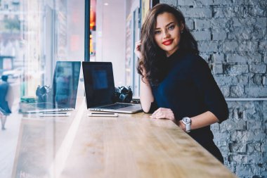 Gülümseyen serbest çalışan, modaya uygun kıyafetler giyen, uzun siyah saçlara dokunup kameraya bakarken akıllı telefon ve laptopla masada duran genç bir kadın fotoğrafçı.