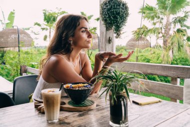 Avuç içleri kalkık, bir kase lezzetli yemek ve bir bardak latte ile masada oturan huzurlu etnik kadın manzarası