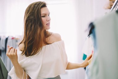 Beyaz bluz giymiş, elbise askılarının yanında duran ve modern mağazayı ziyaret ederken giyinmeyi seçen yoğun kadın.