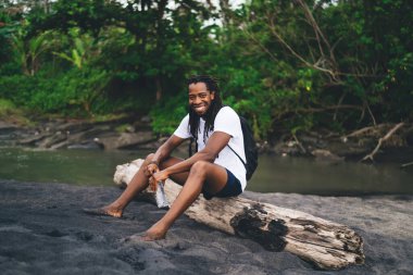 Çıplak ayaklı mutlu Afro-Amerikan erkek. Günlük kıyafetleriyle ağaç gövdesinde oturuyor ve kameraya bakarken gülümsüyor.