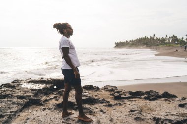 Afrika kökenli Amerikalı bir erkeğin günlük kıyafetleri ve kulaklıkları kumlu sahilde dalgalanan denize yakın duruyor ve gözlerini kaçırıyor.