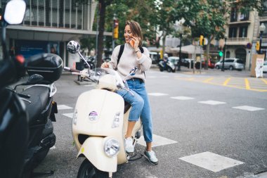 Sırt çantalı, neşeli, yetişkin bir kadın. Scooter 'a binip akıllı telefondan konuşurken gülümsüyor ve başka tarafa bakıyor.