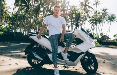 Gündelik giysiler içinde, kumlu tropikal sahilde güneşli yaz saatlerinde avuçlarıyla modern motorsiklete yaslanan ciddi kendine güvenen bir adam.