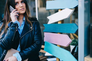Sıradan giysiler içinde, renkli tabelaların yanında dikilip cep telefonuyla konuşan çekici bir kadın.