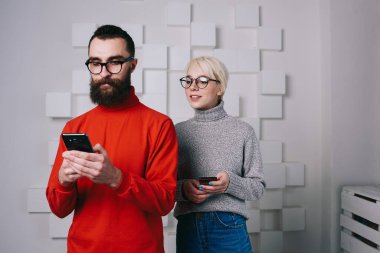 Gri süveterli ve gözlüklü genç sarışın kadın, Geometrik duvarlı genç sakallı hippinin akıllı telefonundan omuz üstünden röntgenliyor.