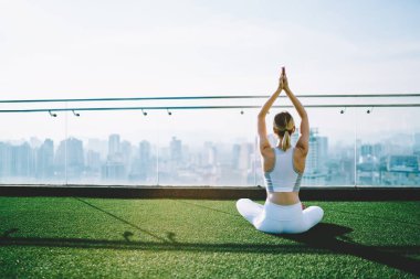 Beyaz spor kıyafetli, Sukhasana yapan, güneşli bir günde büyük bir binanın tepesinde ellerini kaldırmış genç bir kadının arka görüntüsü.