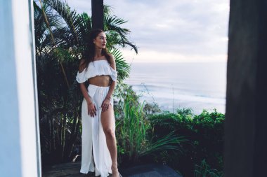 İnce, şehvetli, çıplak ayaklı, şık elbiseli bir kadın beyaz duvarın yanında dikilip günbatımında yaz tatilinin tadını çıkarıyor.