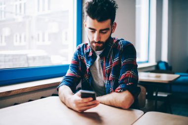 Gündelik kıyafet giymiş sakallı hippi üniversitede masaüstünde oturan ücretsiz 4G internet bağlantısını kullanarak modern akıllı telefondan online mesajlaşma yapıyor. Sosyal ağlarda öğrenci güncelleme profili.