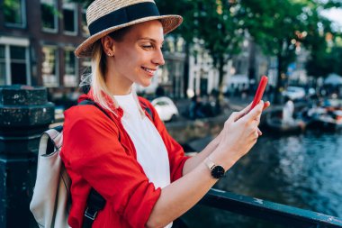 Sırt çantalı, köprüde duran ve Amsterdam 'ı keşfederken cep telefonunu karıştıran pozitif kadın gezginin yan görüntüsü