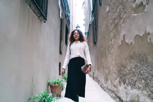 Alçak açılı, kıvırcık saçlı, şık elbiseli, tarihi şehirde seyahat ederken dar şehir caddesinde yürüyen Afro-Amerikan genç bir kadın.