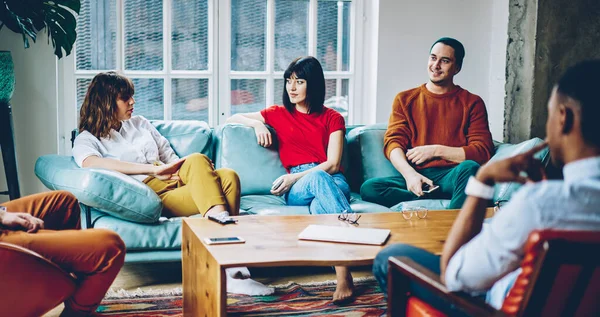 Şık giyinmiş genç bir kadın, sıcak oturma odasındaki kanepenin yanında oturan ilgili bir erkek ve kadınla konuşuyor.