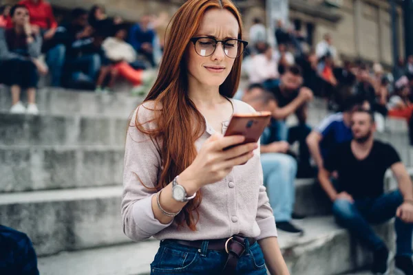 Puzzled female blogger in optical eyewear checking received email message with travel checklist using smartphone device with 4g internet connection