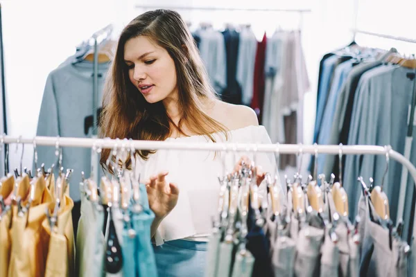 Focused woman in casual dress standing near clothes rails and choosing apparel while spending time in fashion store at daytime
