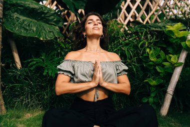 Moda kıyafetleri içinde mutlu genç etnik bayan gezgin. Tropikal bir ülkede yaz tatilinde Lotus pozunda namaste hareketleriyle yoga meditasyonu yapıyor.