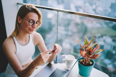 Masada dizüstü bilgisayarla oturan ve balkonda içki içen genç kadın kartviziti yüksek açılı.