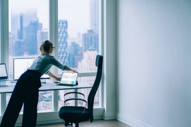 Resmi giyim ve gözlük takan meşgul bayanın arka planı veri üzerinde çalışırken tableti diğer cihazların yanına yerleştiriyor.