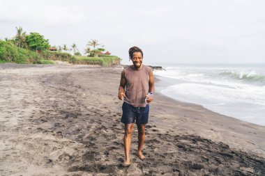 Kumlu tropikal deniz kıyısında dururken elinde bir şişe suyla gülümseyen yazlık elbiseli mutlu Afro-Amerikalı bir adam.