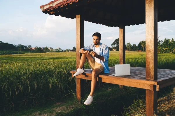 Full body focused on screen of smartphone man in casual outfit working on laptop remotely while spending time on wooden terrace on nature