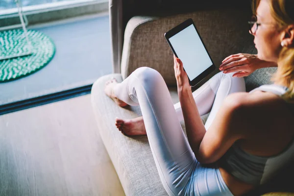 Back view of young blond female in white sport outfit and eyeglasses reading book on tablet while relaxing on cozy sofa in apartment