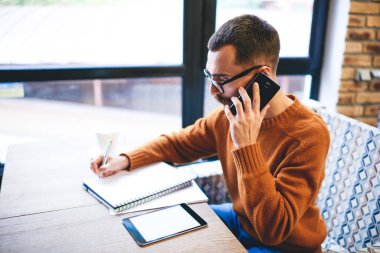İş sırasında masada oturup notlar yazan ve cep telefonuyla konuşan ciddi erkek yüksek açılı.