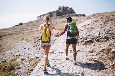 Yarı çölde yüzsüz erkek ve dişinin sırt çantalarıyla arka planda eski bir binayla birlikte yürüdüğünü gösteren tüm vücut görüntüsü.