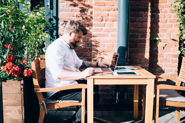 天気の良い日にリモートで作業しながら 屋外カフェで木製の椅子に座っている集中男性マネージャーブラウジングネットブックの側面図 — ストック写真