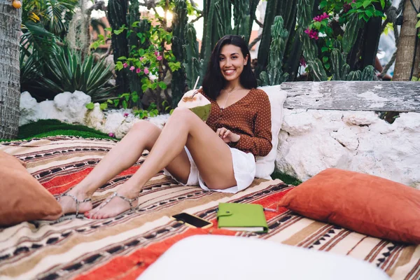 Cheerful young ethnic lady in summer clothes enjoying coconut water in green shell and leaning on pillow while sitting on patterned rug