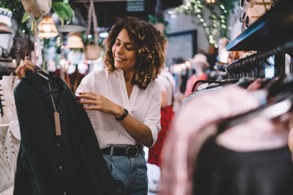 Smiling female standing near hangers with brand wear and laughing during Black Friday shopping with discounts