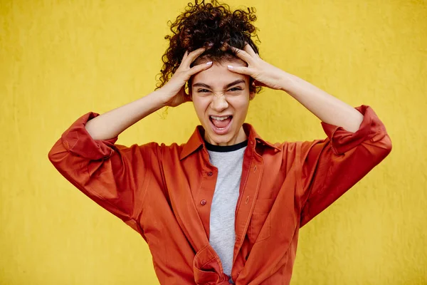 Furious ethnic female in casual outfit with curly hair looking at camera and opening mouth while shouting and holding hands on head