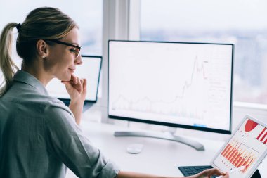 Kadın finansal analistin çenesine dokunurken iş yerindeki masaüstü bilgisayarının yanında tablet ekranına Stylus 'u işaret ederken kenar görünümü