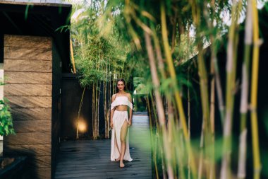 Yeşil bahçenin süslü girişinin yanında duran ve kameraya bakan, beyaz giysili, tanınmayan, çıplak ayaklı bir kadın.