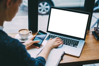 Yukarıdan, akıllı telefon ve dizüstü bilgisayar kullanırken bir fincan kahve ve planlayıcıyla masada oturan serbest çalışan kadın.