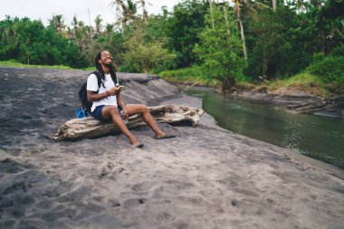 Nehir kıyısındaki ağaç gövdesinde otururken akıllı telefondan gülümseyen ve mesajlaşan gündelik kıyafetlerle uzun boylu mutlu Afrikalı Amerikalı bir erkek.