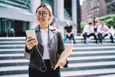 Gözlüklü ve kulaklıklı neşeli hippi kızın yarım boy portresi pozitif müzik şarkıları dinliyor ve şehirde vakit geçirirken kameraya gülümsüyor.