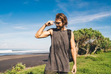 Positive man in sunglasses with backpack standing alone near waving sea while resting on summer day on tropical beach
